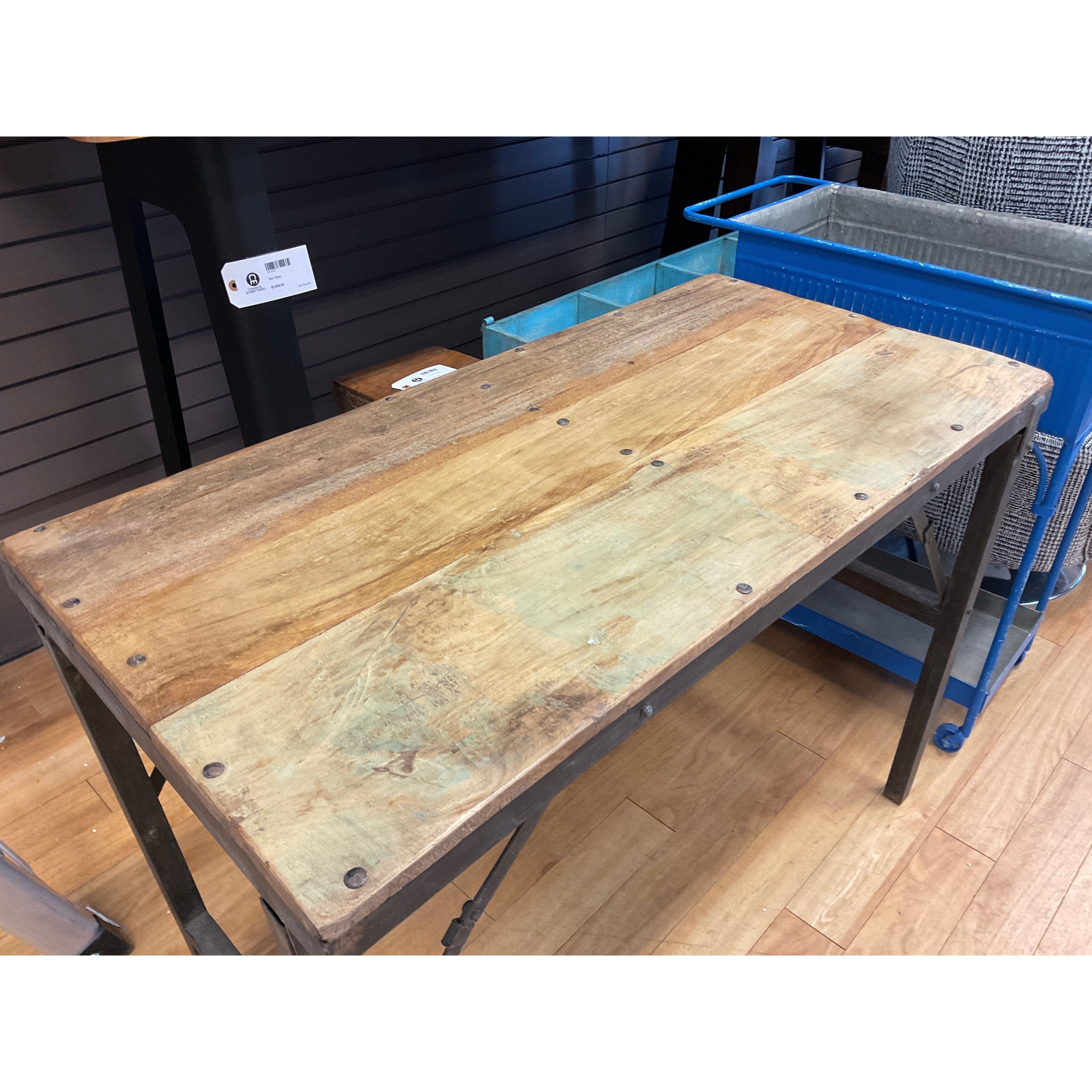 The Wedding Table features a hand-crafted rectangular wooden top with visible grain and natural imperfections, set on a metal frame. Metal rivets accent the corners, enhancing its unique artisanal style, perfect for any light wood floor setting.