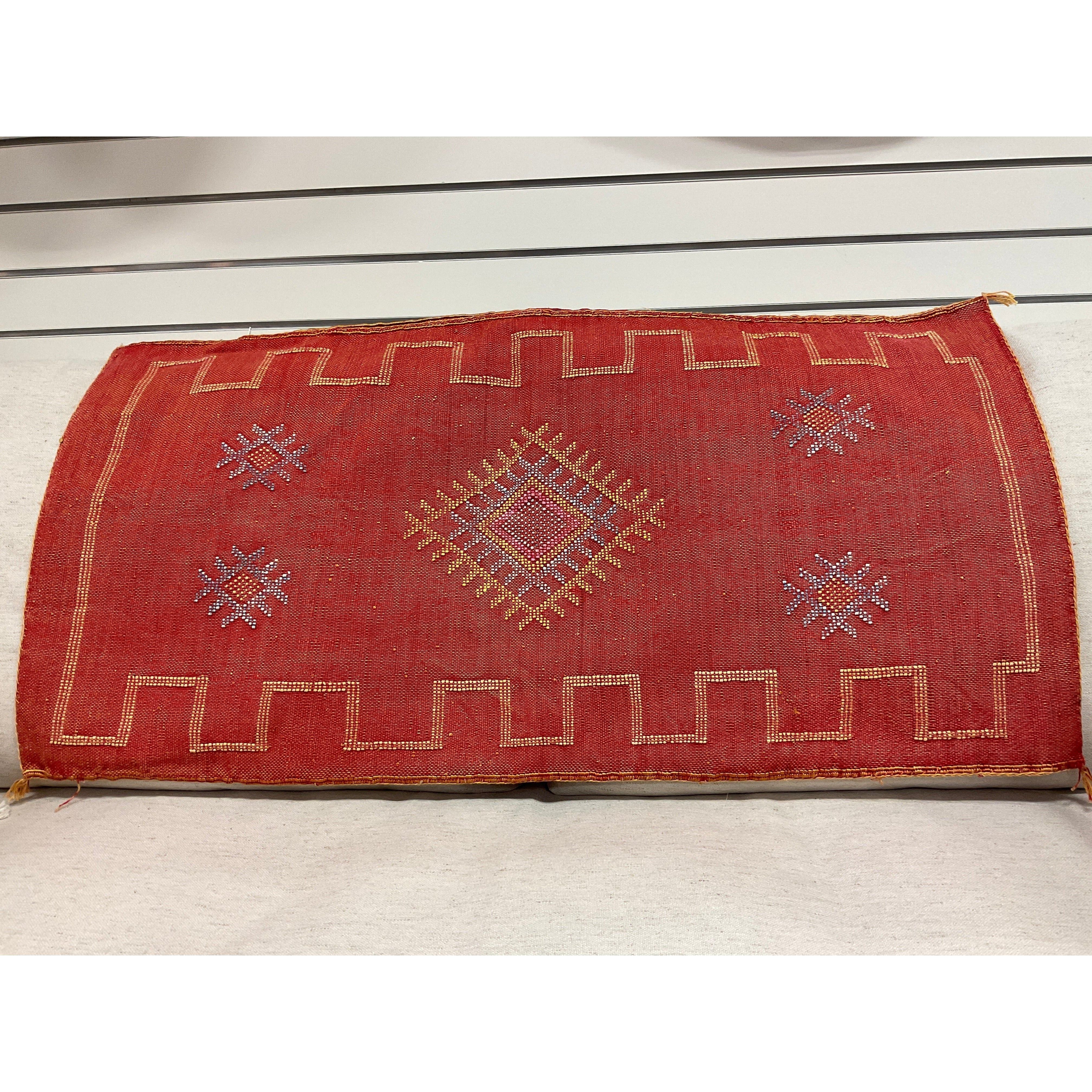 A rectangular red woven textile with a symmetrical design highlights an intricate weaving technique. The center features a diamond pattern in yellow and white, surrounded by smaller geometric shapes. It boasts a decorative border along the edges. The white background with horizontal lines suggests it's displayed on a wall.

Carnelian Cactus Fiber Throw Pillow