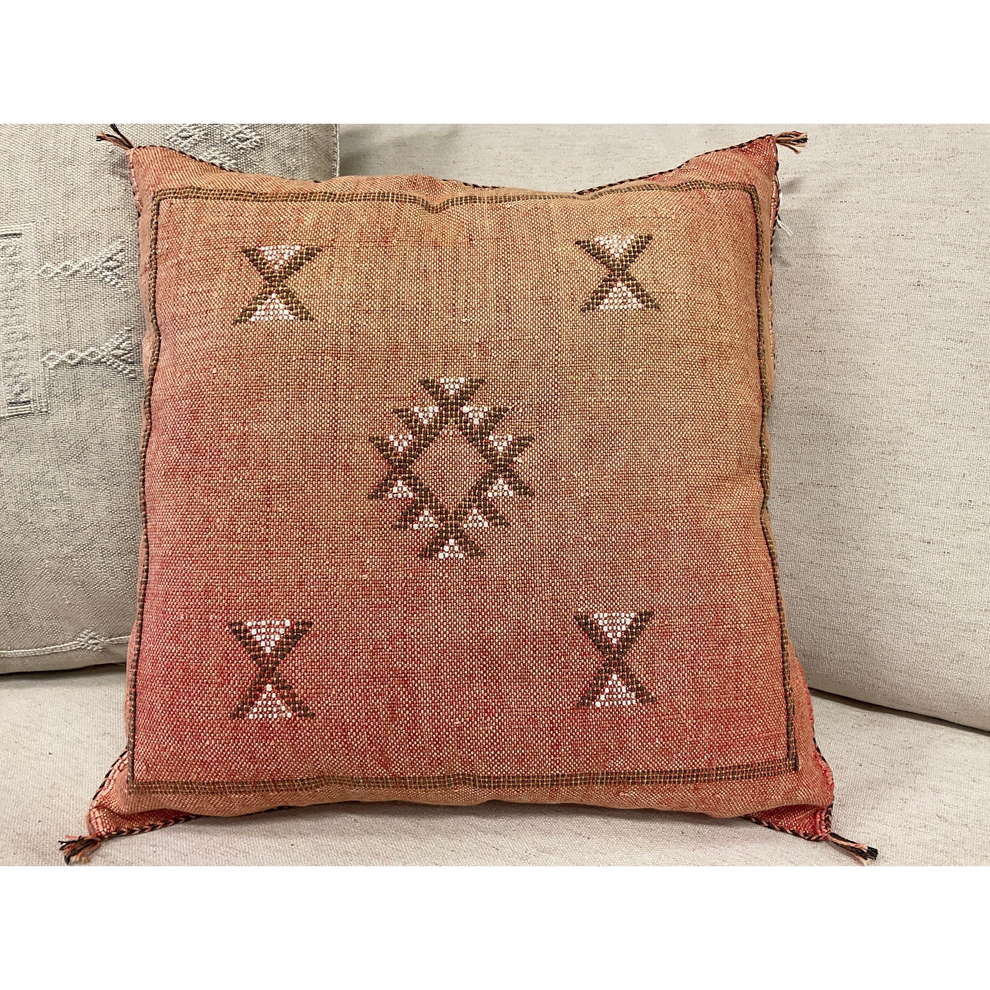 A Carnelian Cactus Fiber Throw Pillow with a faded reddish-orange hue featuring a diamond pattern in the center, flanked by two X-shaped symbols above and below. The Carnelian Cactus Fiber Throw Pillow has frayed corners and is placed on a light gray sofa. A portion of another cushion with an intricate weaving technique is visible on the left.