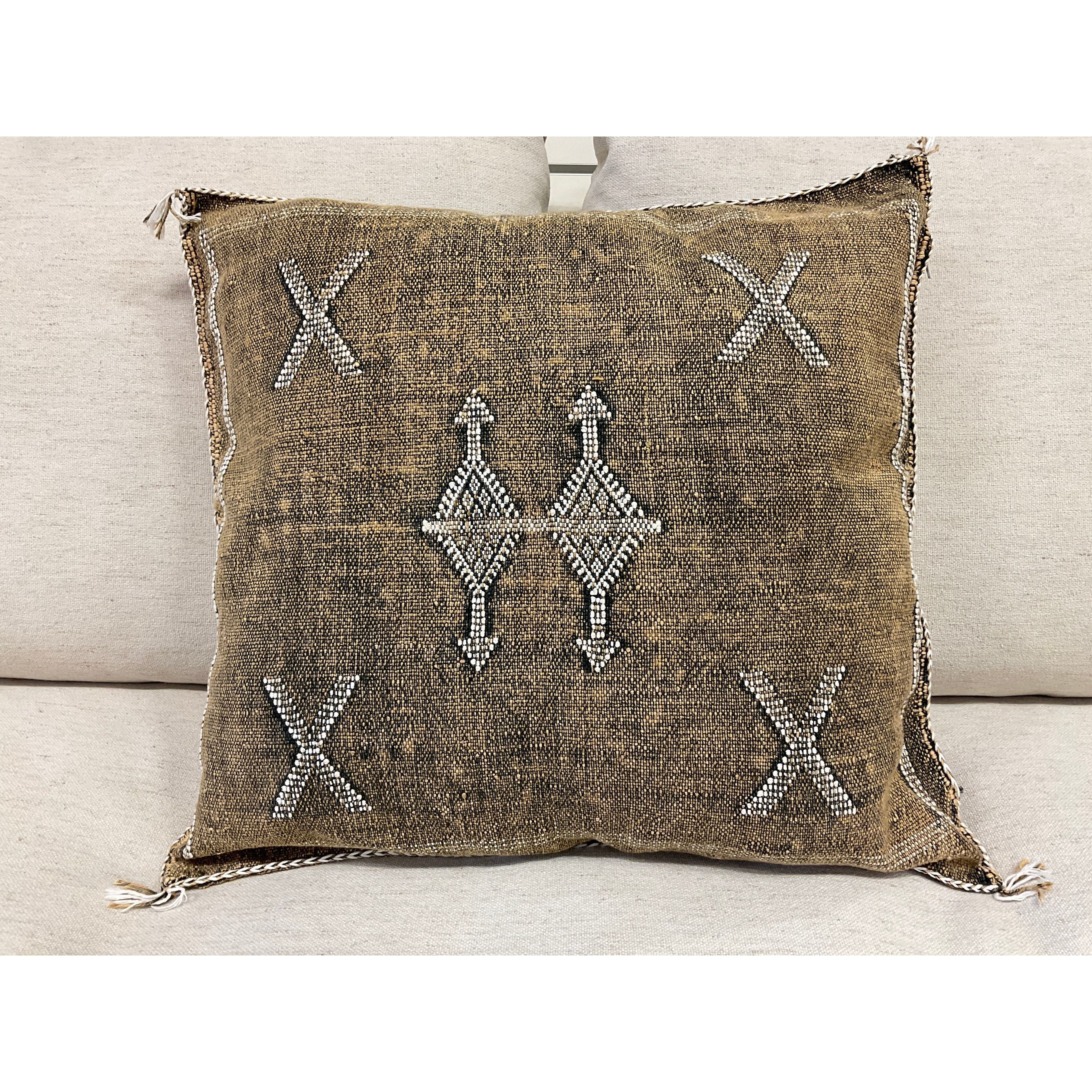 The Earth Cactus Fiber Throw Pillow is a square cushion with a brown fabric cover and white geometric embroidery. It features four corner crosses, a central diamond of interlocking lines, frayed edges, and tassels on the corners against a light grey background.