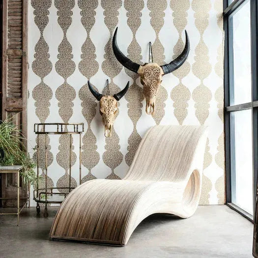 A modern wooden chaise lounge sits by a window, with two Carved Lace Skull Wall Art pieces featuring ornate carvings and dark horns mounted above. A metal side table and green plant complete the space against gold-patterned wallpaper.