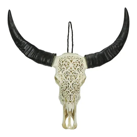 The Carved Lace Skull Wall Art features a handcrafted cow skull with large dark horns, showcasing intricate floral and scrollwork patterns. It comes with a black hanging cord for display against any plain background.