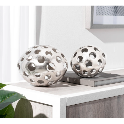 Two Sandcast Orb sculptures, featuring a metallic finish and circular cutouts, are crafted from aluminum. They rest on a light wooden console, with one orb being larger than the other. In the background is a white curtain alongside framed wall art. The minimalistic decor is beautifully enhanced by natural light creating soft shadows, while a leaf gently peeks in from the bottom left corner.