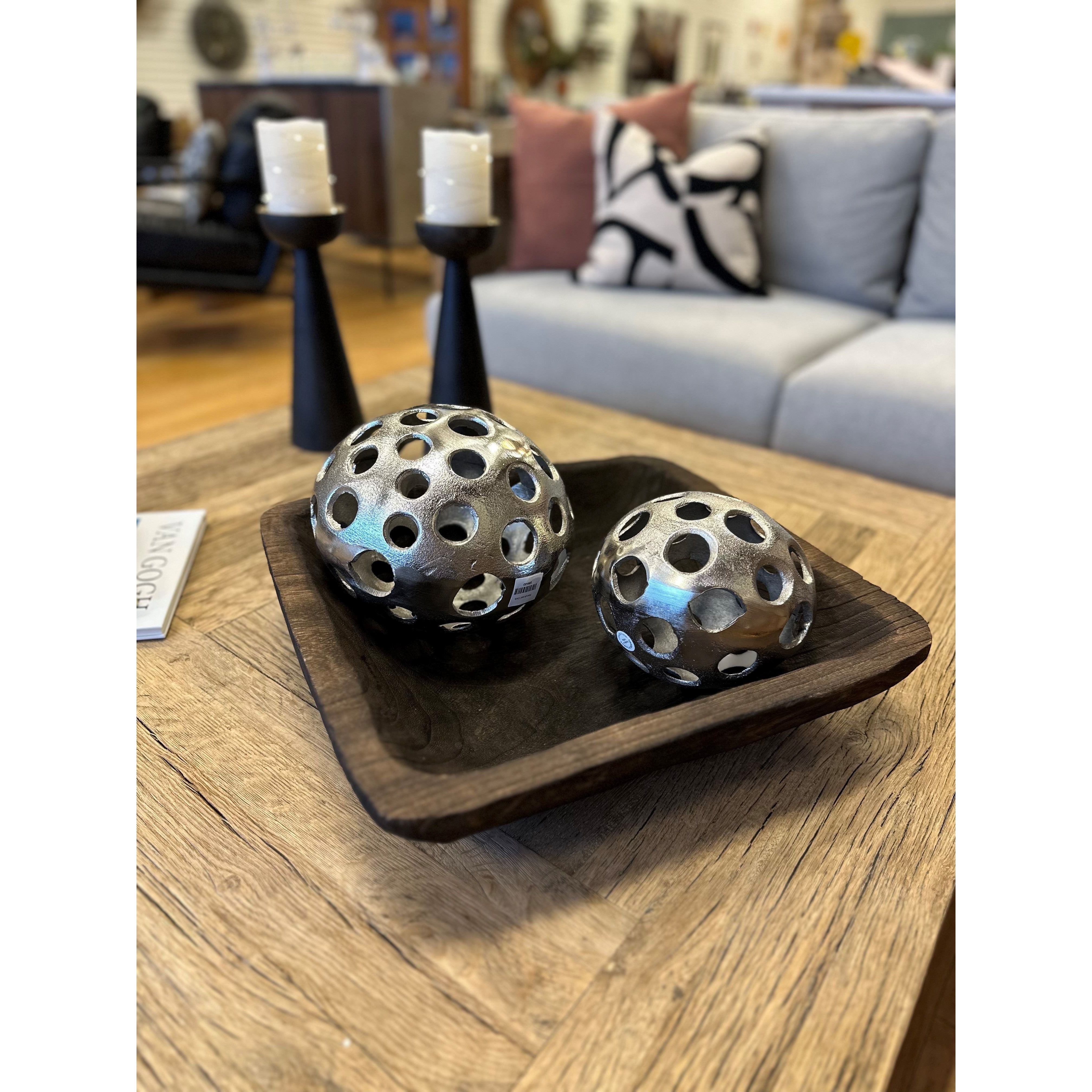 A Paulownia Display Trough sits on the coffee table, holding two silver sphere sculptures with circular cutouts. Two black candleholders with white candles are behind it, in front of a gray sofa with patterned pillows in a cozy living room.