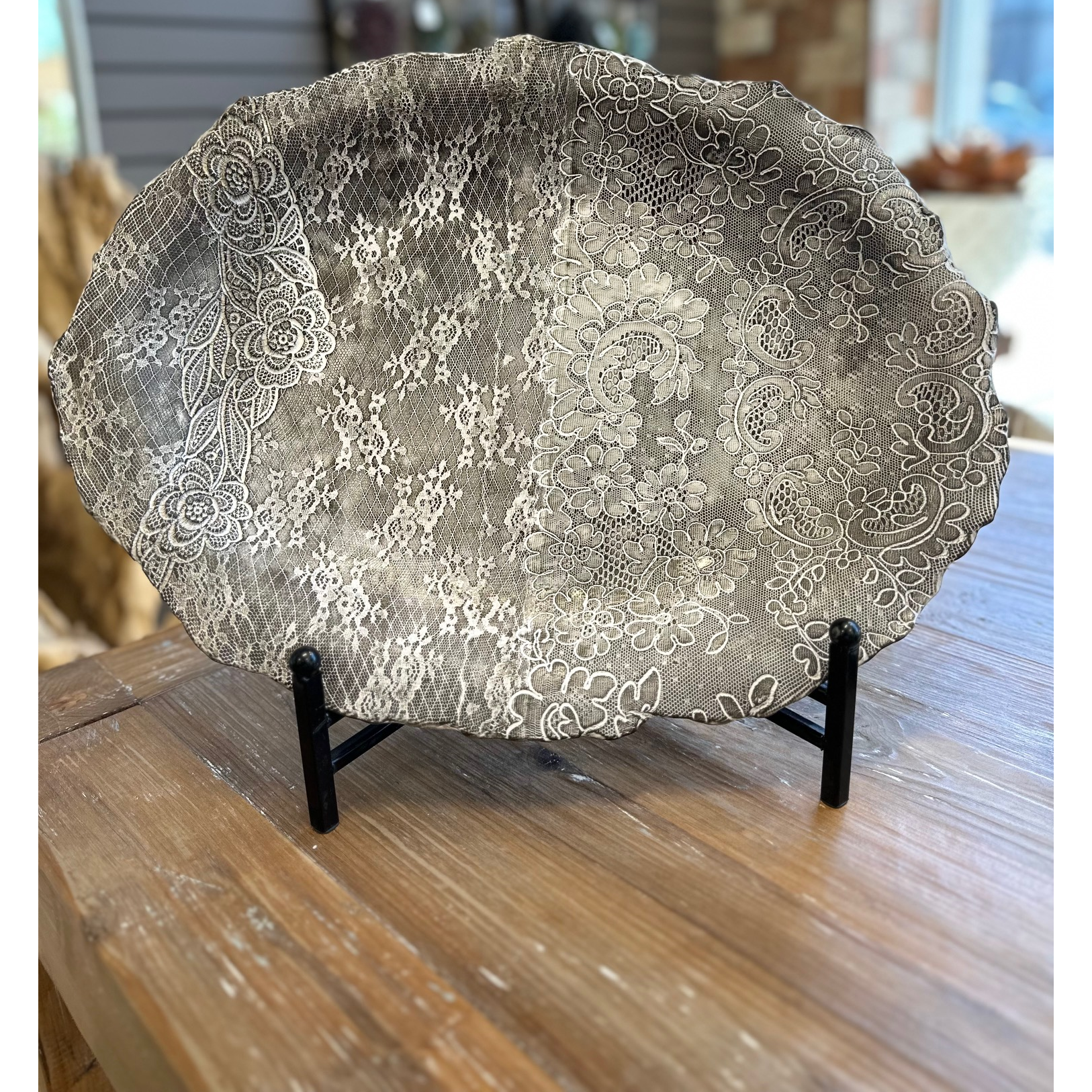 The Casado Serving Platter, crafted by a French ceramist, showcases delicate white floral lace patterns on a gray ceramic base with a scalloped edge, displayed on a black stand atop a wooden table to highlight its artisanal beauty.