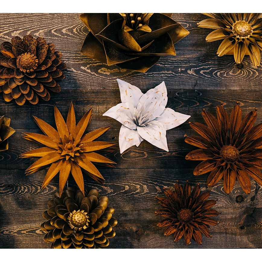 The Rust Aster Metal Flower features brown, gold, and bronze tones arranged on dark wood with a standout white center, creating striking wall-mounted decor ideal for outdoor spaces.