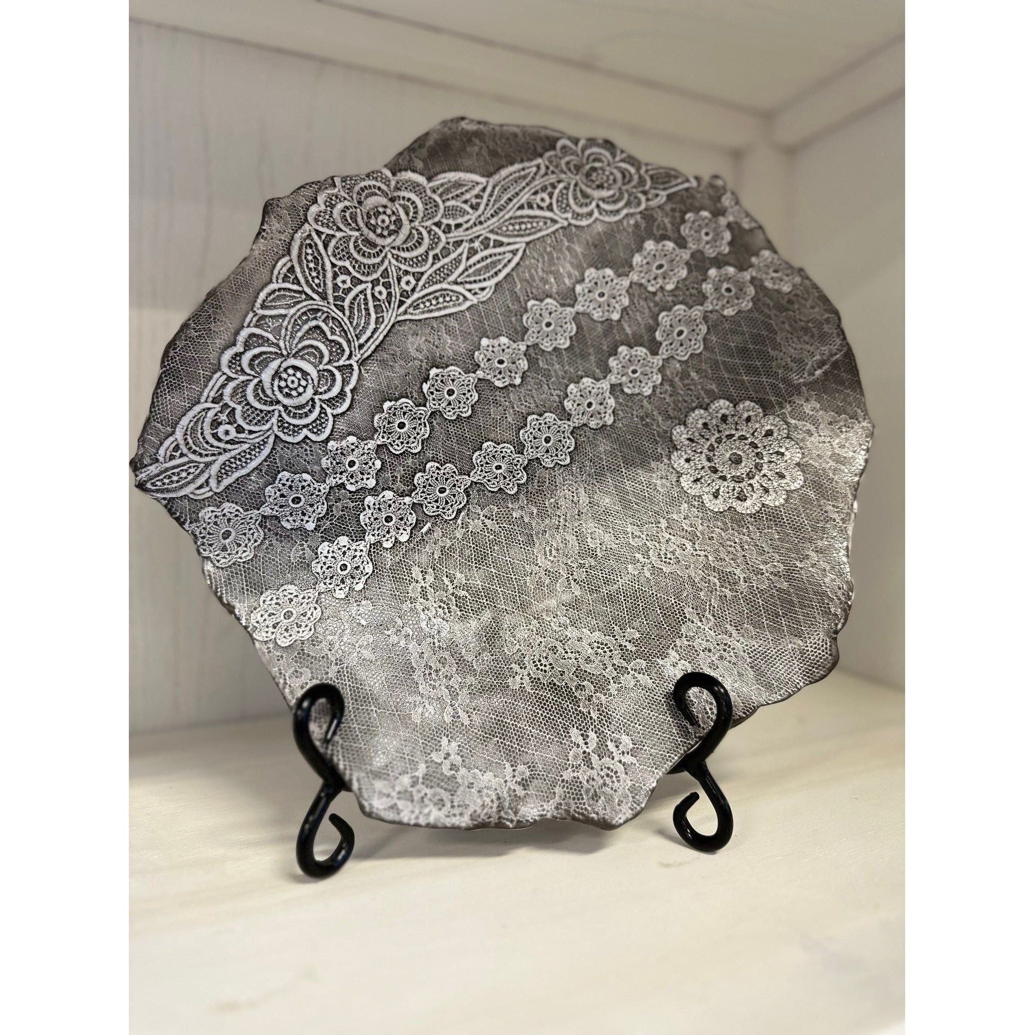 The Casado Serving Platter, a round ceramic piece with uneven edges by a French ceramist, features intricate white lace and floral designs. Handcrafted and set on a black metal stand, it adds vintage charm to any light wooden shelf.