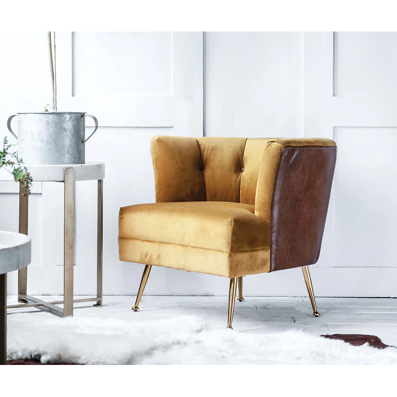 The Lobby Accent Chair features channel tufting with mustard yellow velvet front and brown leather back, set on brass legs. It sits in a bright room with a white fur rug, side table, metal watering can, and minimal decor.